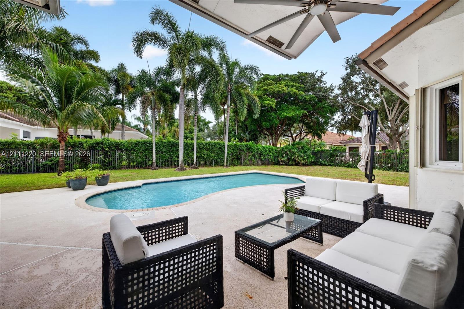 Covered patio featuring an integrated ceiling fan, concrete deck, kidney-shaped swimming pool, lush perimeter hedge, and mature palm trees