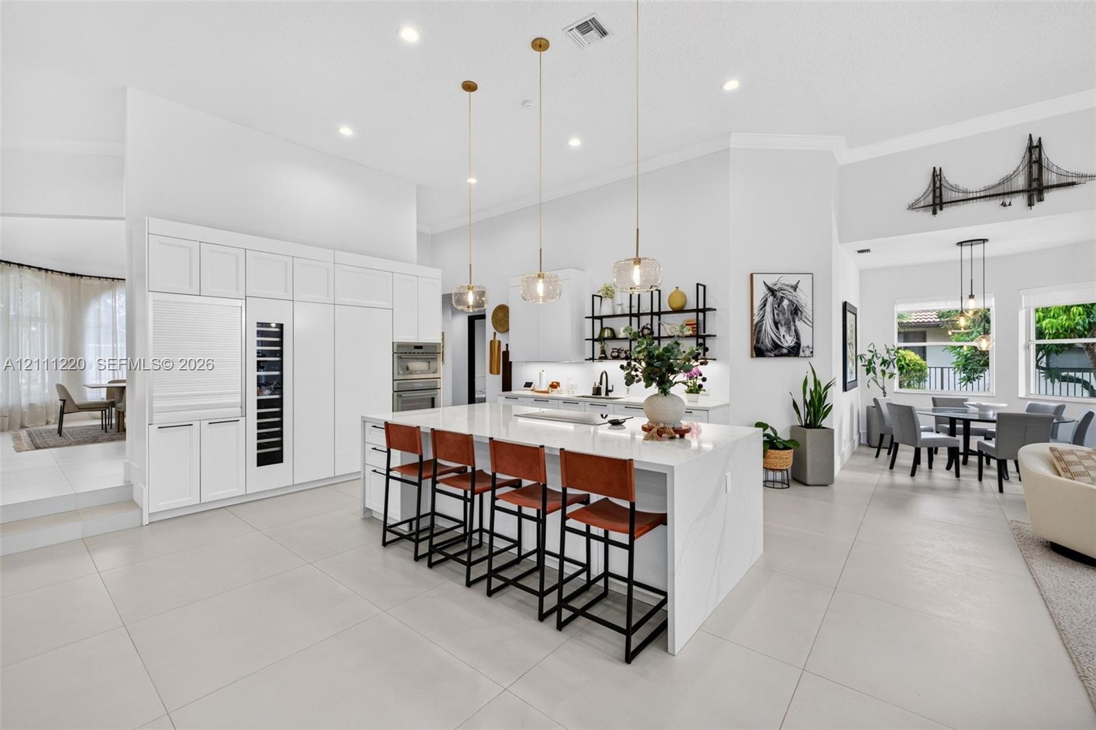 Gourmet kitchen featuring a large central island with stone-finish countertops, custom floor-to-ceiling white cabinetry, integrated stainless steel appliances, tiered pendant lighting, and oversized neutral tile flooring