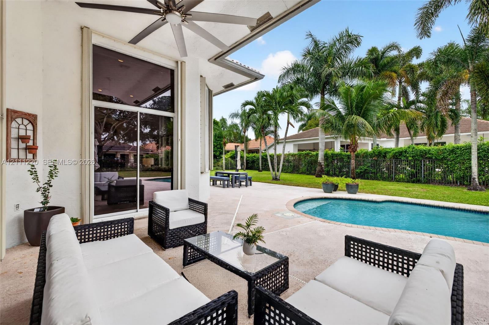 Covered patio featuring a ceiling fan, sliding glass doors, concrete decking, and a swimming pool set within a fenced yard with mature palms