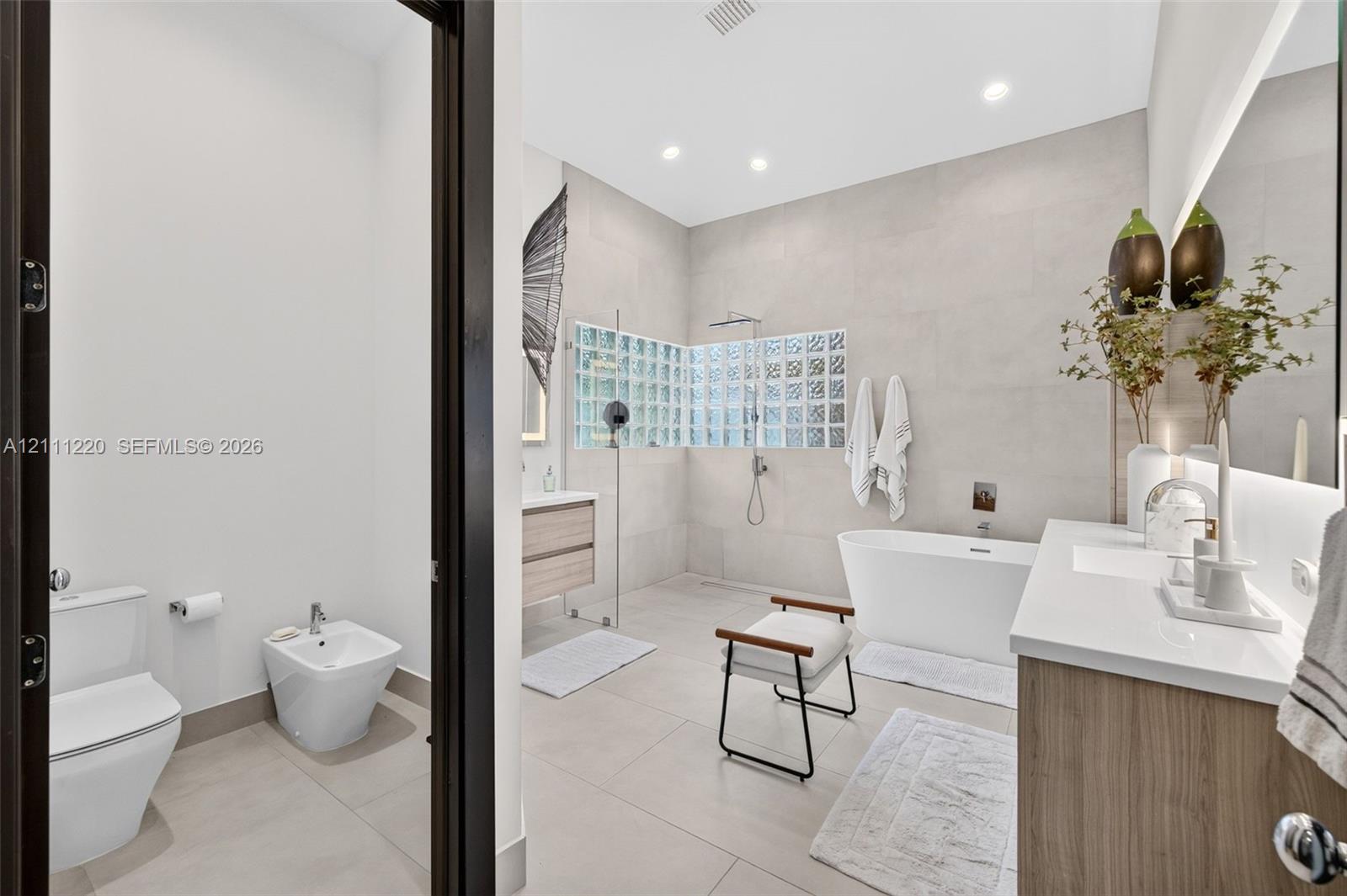 Bathroom featuring a large freestanding soaking tub, walk-in shower with glass block accent wall, double vanity with wood-finish cabinetry, wall-mounted faucet fixtures, and ceramic tile flooring