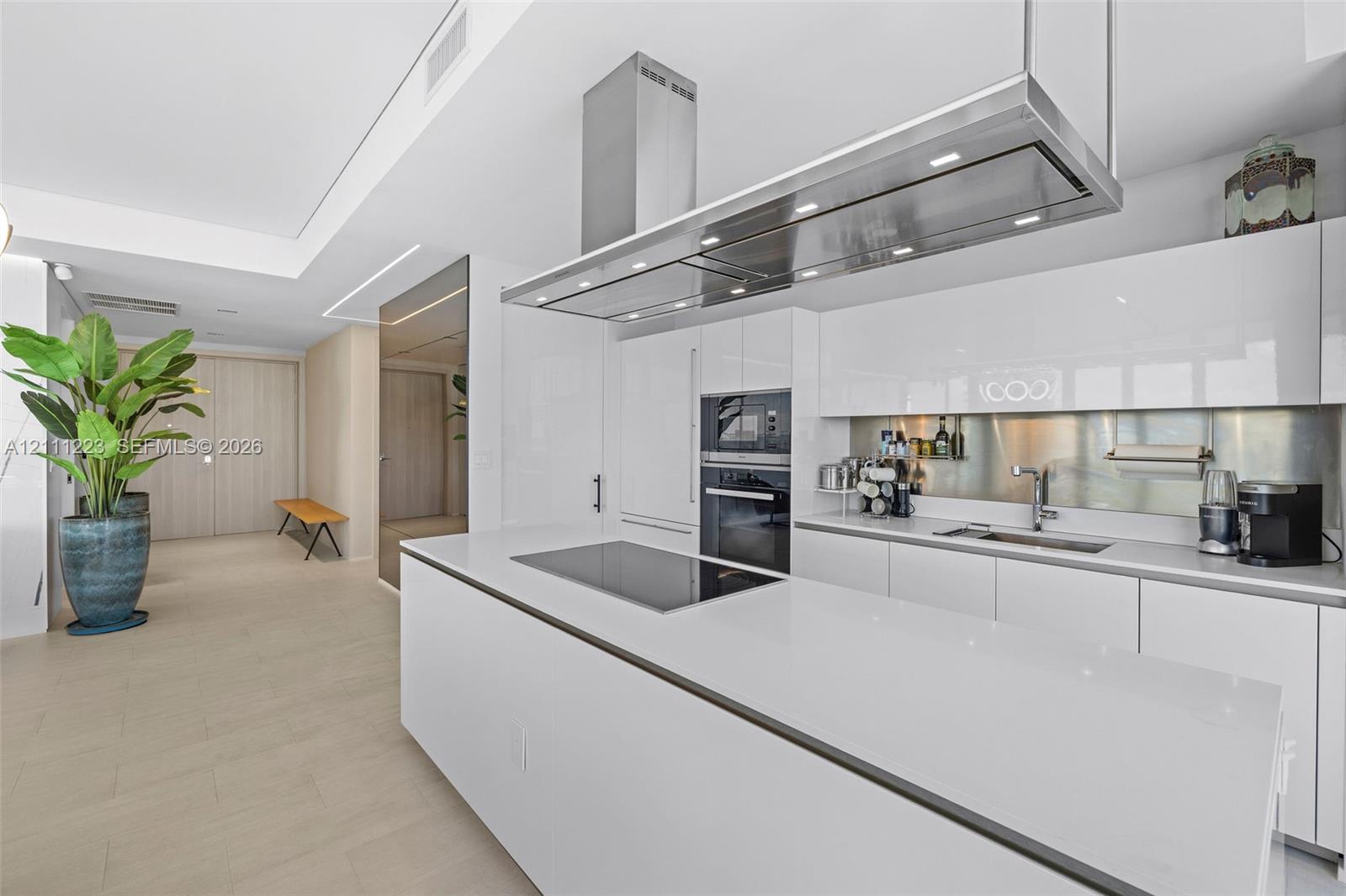 Kitchen featuring high-gloss cabinetry, a spacious island with an integrated induction cooktop, a stainless steel range hood, and a metallic backsplash