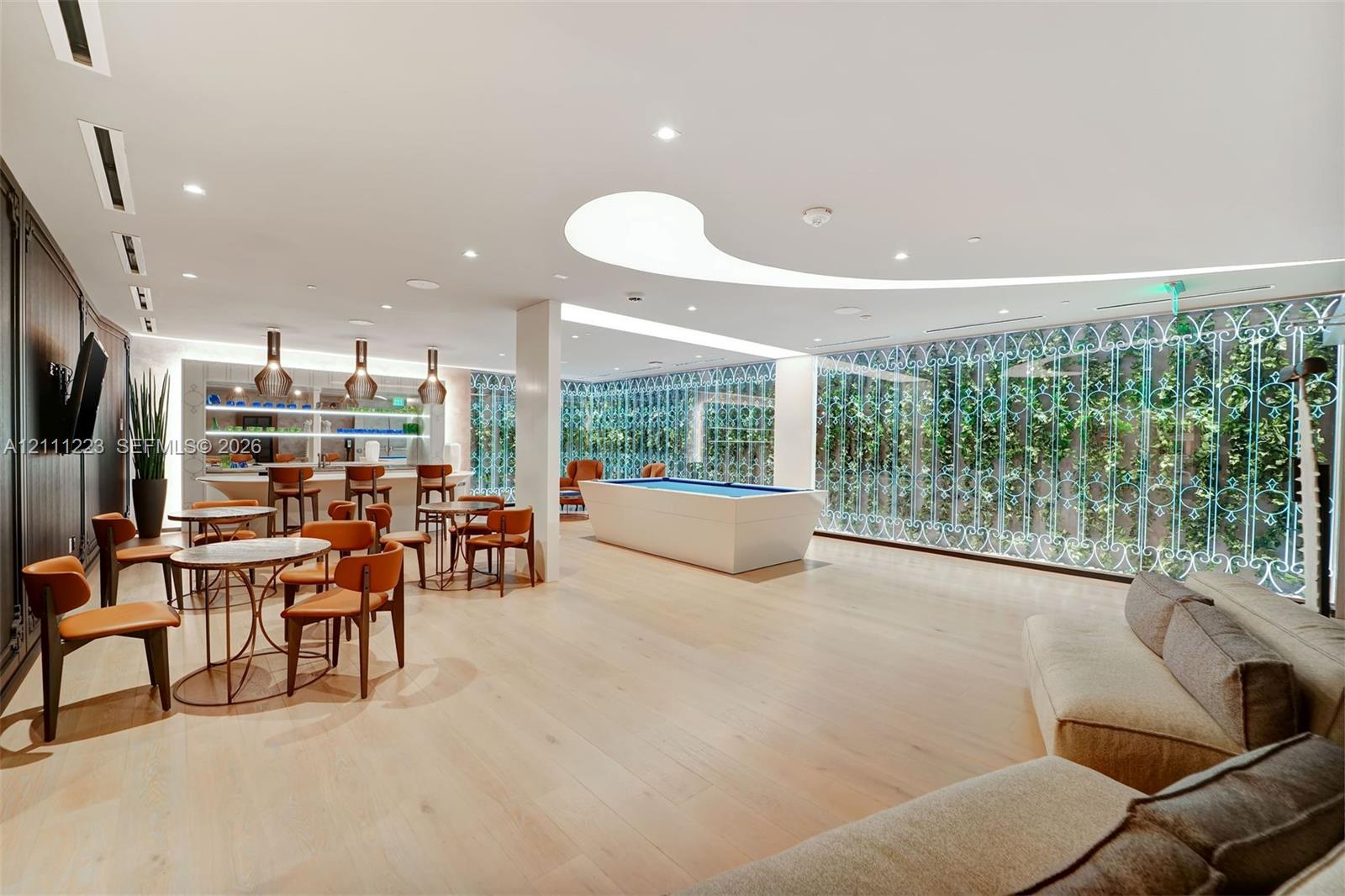 Large common area featuring light-toned wood-finish flooring, a curved ceiling cove light fixture, built-in bar with pendant lighting, and expansive floor-to-ceiling lattice screen windows