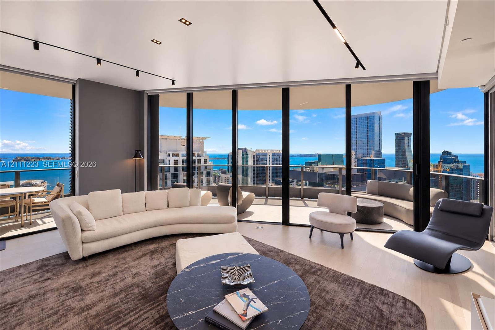 Floor-to-ceiling glass walls, curved balcony with railing, light-tone flooring, recessed track lighting, and panoramic urban and water scenery