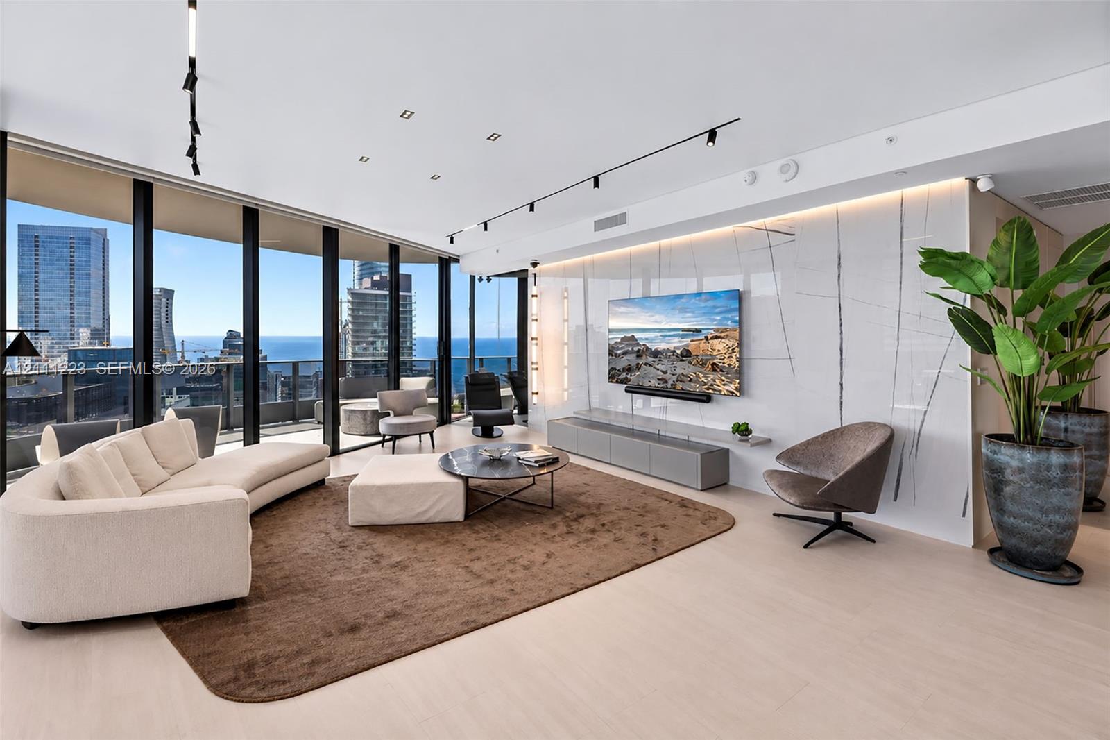 Floor-to-ceiling windows featuring panoramic city and water scenery, light-toned wood-finish flooring, large-format stone feature wall with integrated recessed lighting, and modern track lighting system