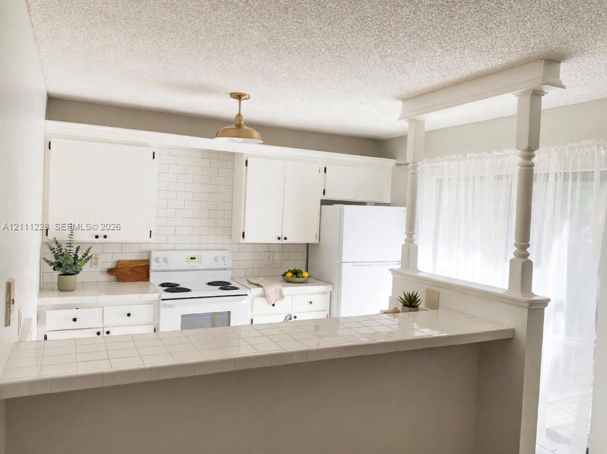 Kitchen featuring white cabinetry, subway tile backsplash, ceramic tile countertop, pass-through breakfast bar with decorative columns, and metallic pendant lighting