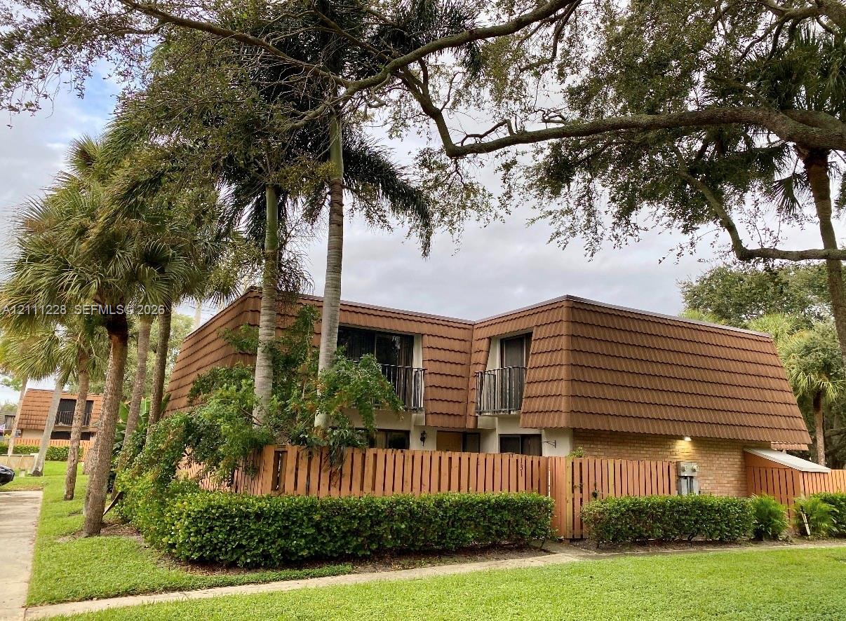 Two-story exterior featuring a mansard roof, brick facade, wood-slat privacy fencing, and second-level balconies shaded by mature palms and canopy trees Two-story exterior featuring a mansard roof, brick facade, wood-slat privacy fencing, and second-level balconies shaded by mature palms and canopy trees