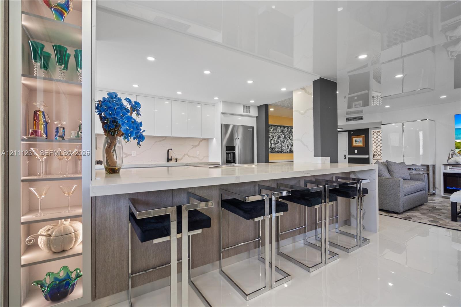 Kitchen featuring gloss-finish cabinetry, stone-surface countertop with waterfall edge, built-in illuminated glass shelving, and stainless steel refrigerator