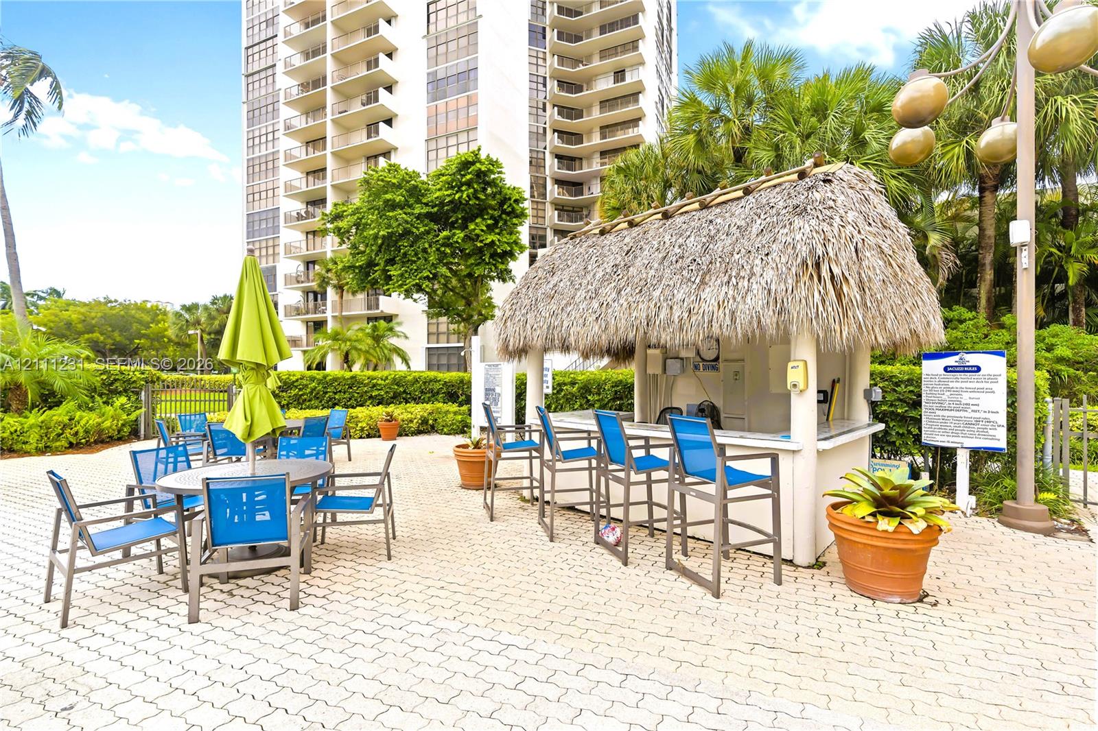 Outdoor poolside bar structure featuring a thatched roof, concrete service counter, paver stone patio, and perimeter hedge landscaping