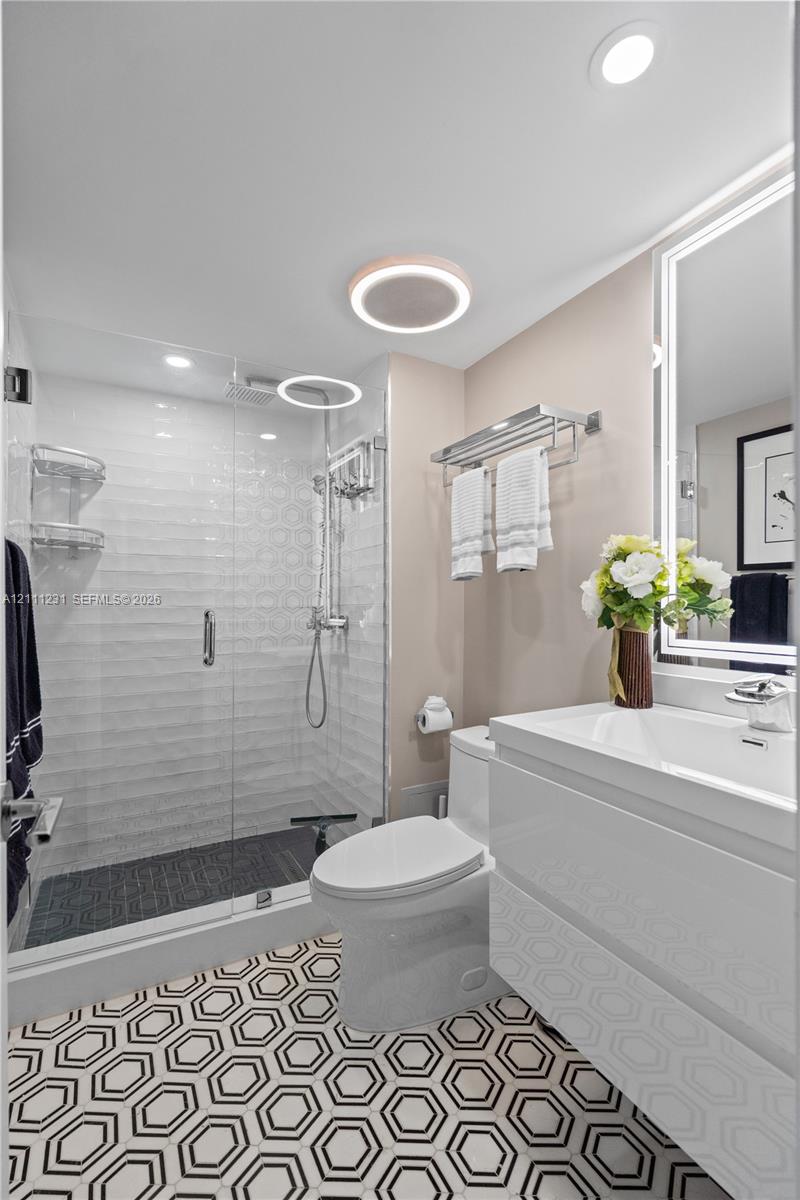 Bathroom featuring a walk-in shower with glass enclosure, wall-mounted rainfall showerhead, geometric patterned floor tiling, white floating vanity, and an integrated LED-backlit mirror