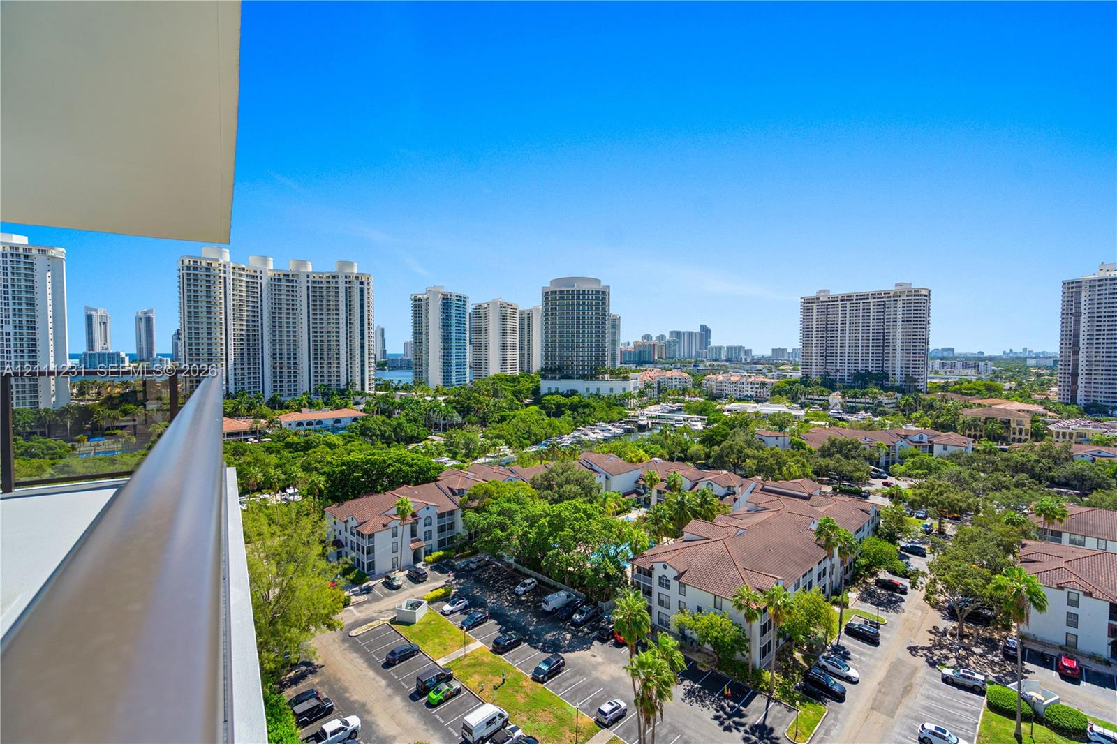Covered balcony featuring a metal railing and expansive urban surroundings including mid-rise residential structures, mature landscaping, and paved parking areas