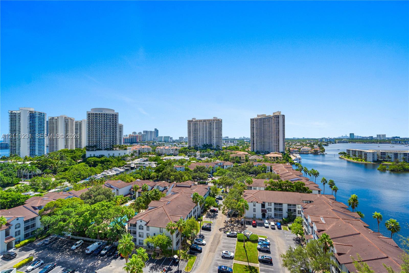 Aerial perspective featuring multi-story residential buildings, paved surface parking, mature palm trees, and waterfront access