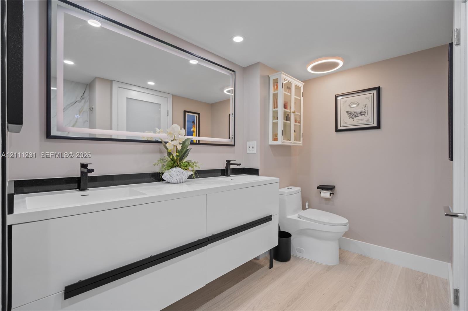 Modern floating vanity featuring dual integrated sinks, matte black faucets, and a dark stone backsplash, paired with a large backlit vanity mirror and built-in wall cabinetry