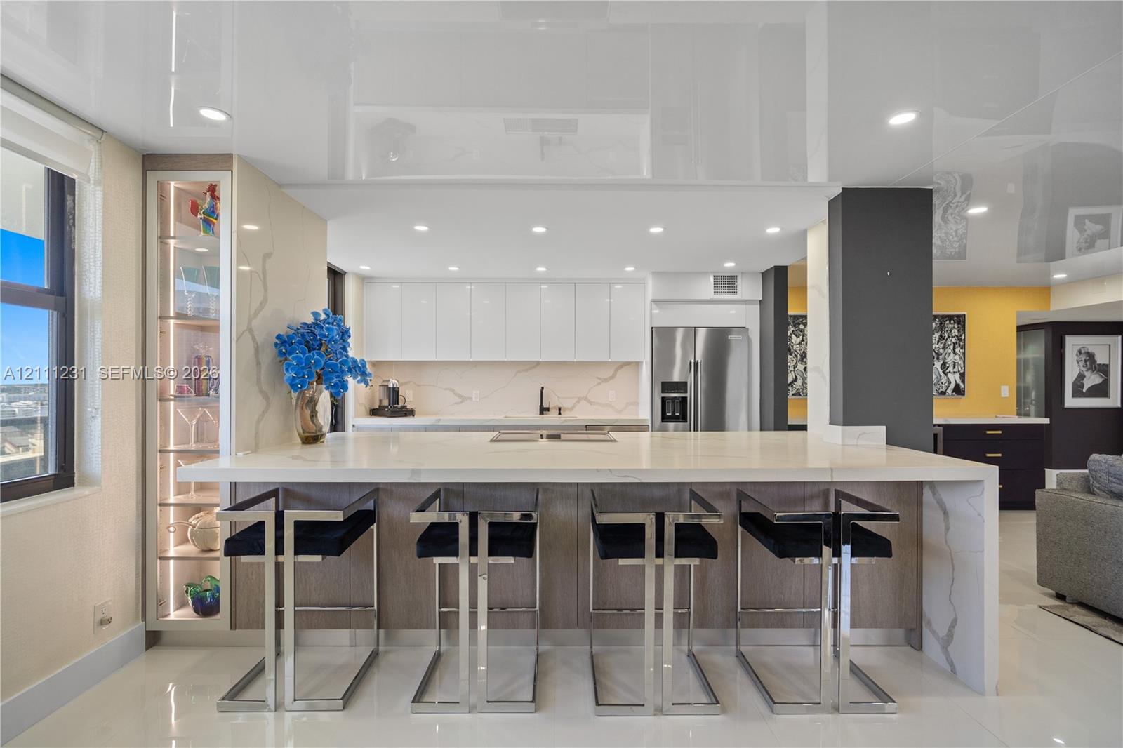 Kitchen featuring a waterfall-edge island with stone-finish countertop, high-gloss ceiling, integrated display cabinetry with interior lighting, sleek flat-panel cabinetry, and stainless steel refrigerator