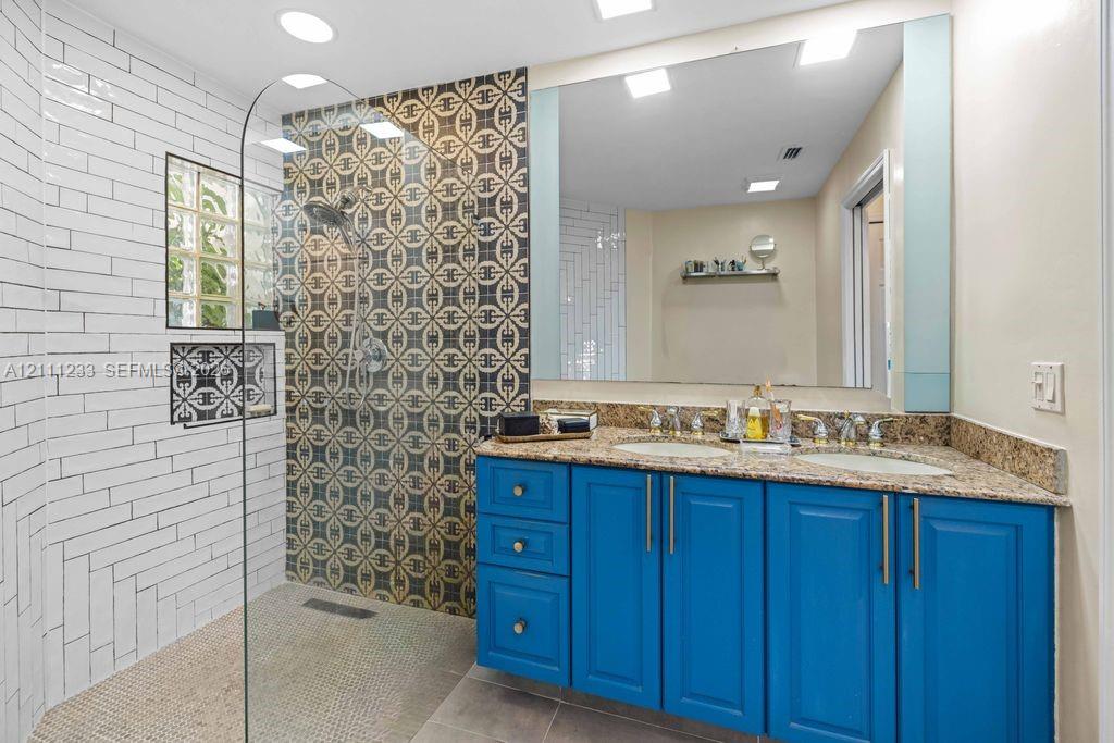 Bathroom featuring a double-sink vanity with stone-finish countertop, walk-in shower with glass panel, patterned tile accent wall, white subway tile surround, and large wall-mounted mirror