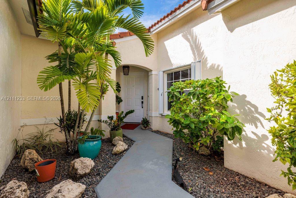 Stucco exterior entrance featuring a concrete walkway, arched entryway, and mature landscaping with rock bed mulch