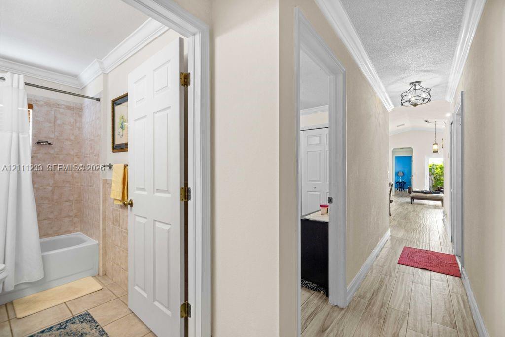 Hallway featuring wood-finish flooring, crown molding, and a multi-arm flush-mount light fixture, with a tiled bathroom including a tub-shower combination