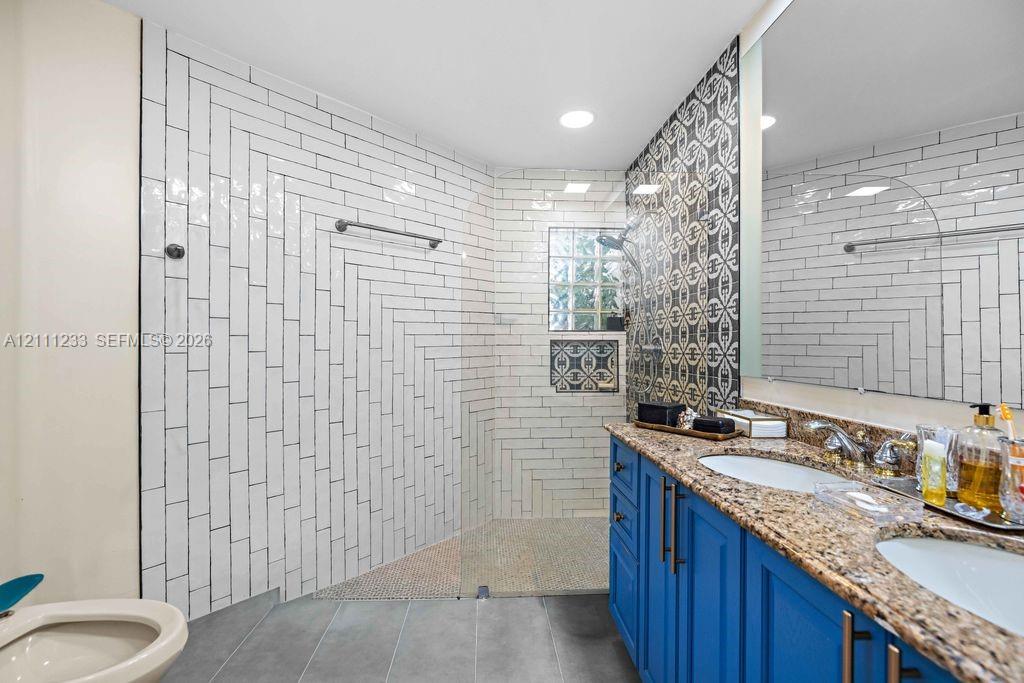 Bathroom featuring a double-sink vanity with stone-surface countertop, walk-in shower with glass panel, herringbone-patterned wall tile, and decorative accent tile work