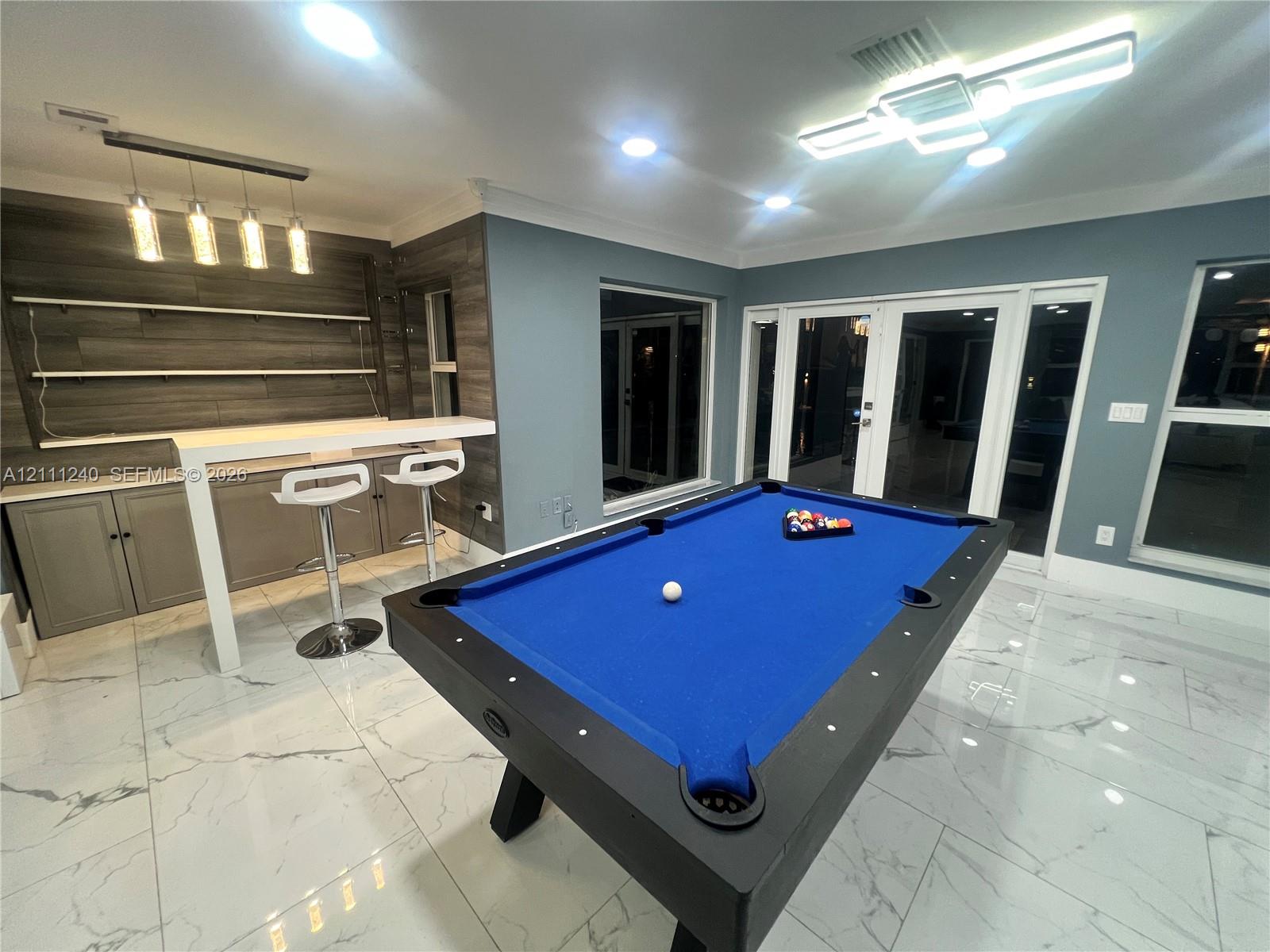 Large-format tile flooring, built-in bar with wood-finish wall paneling, floating shelving, multi-pendant bar lighting, and French doors