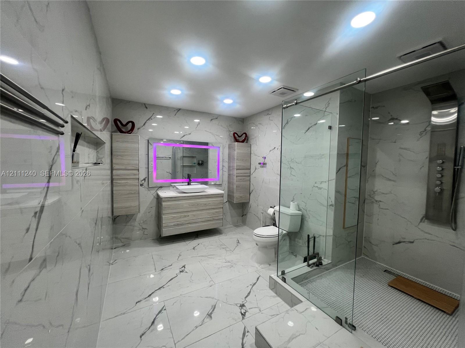 Full-height large-format wall and floor tile, wood-finish floating vanity with vessel sink, glass-enclosed shower featuring a built-in rainfall panel, recessed ceiling lighting, and wall-mounted storage cabinetry