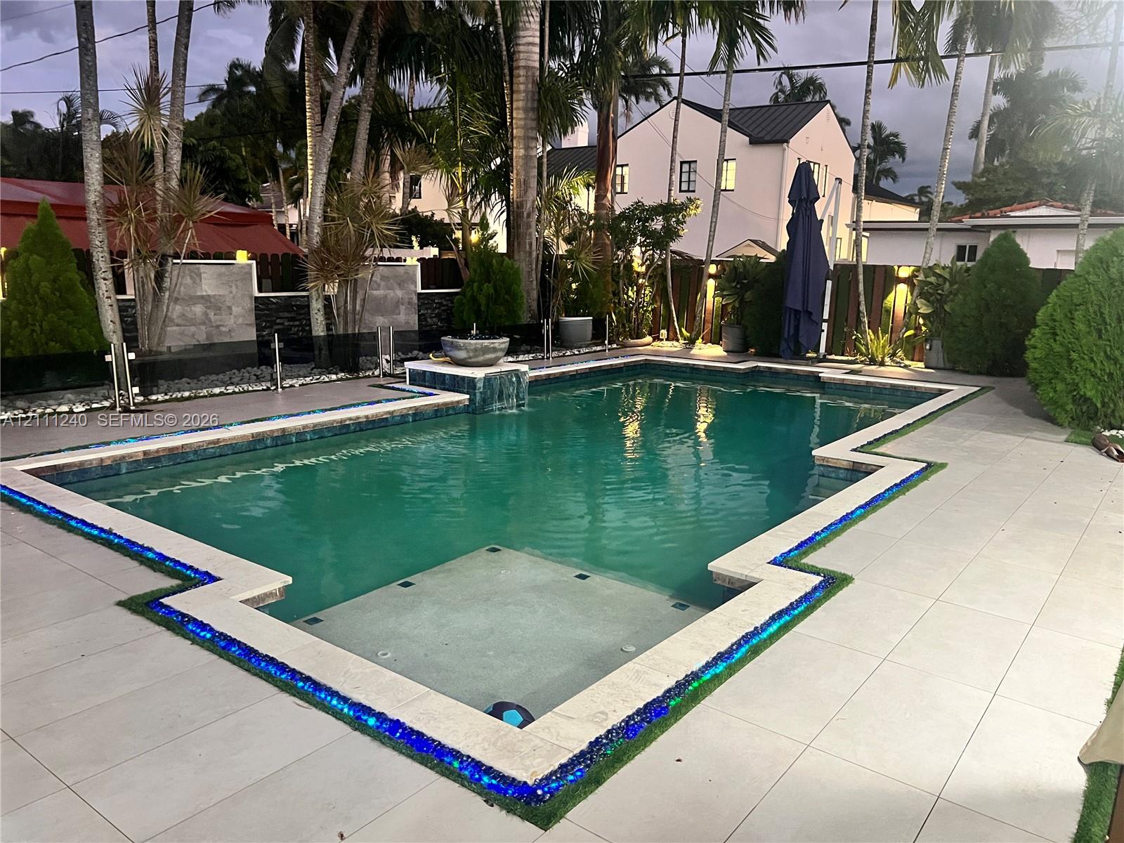 Custom-shaped swimming pool featuring a built-in water feature, mosaic tile perimeter inlay, light-colored tile decking, and stone-surround walling
