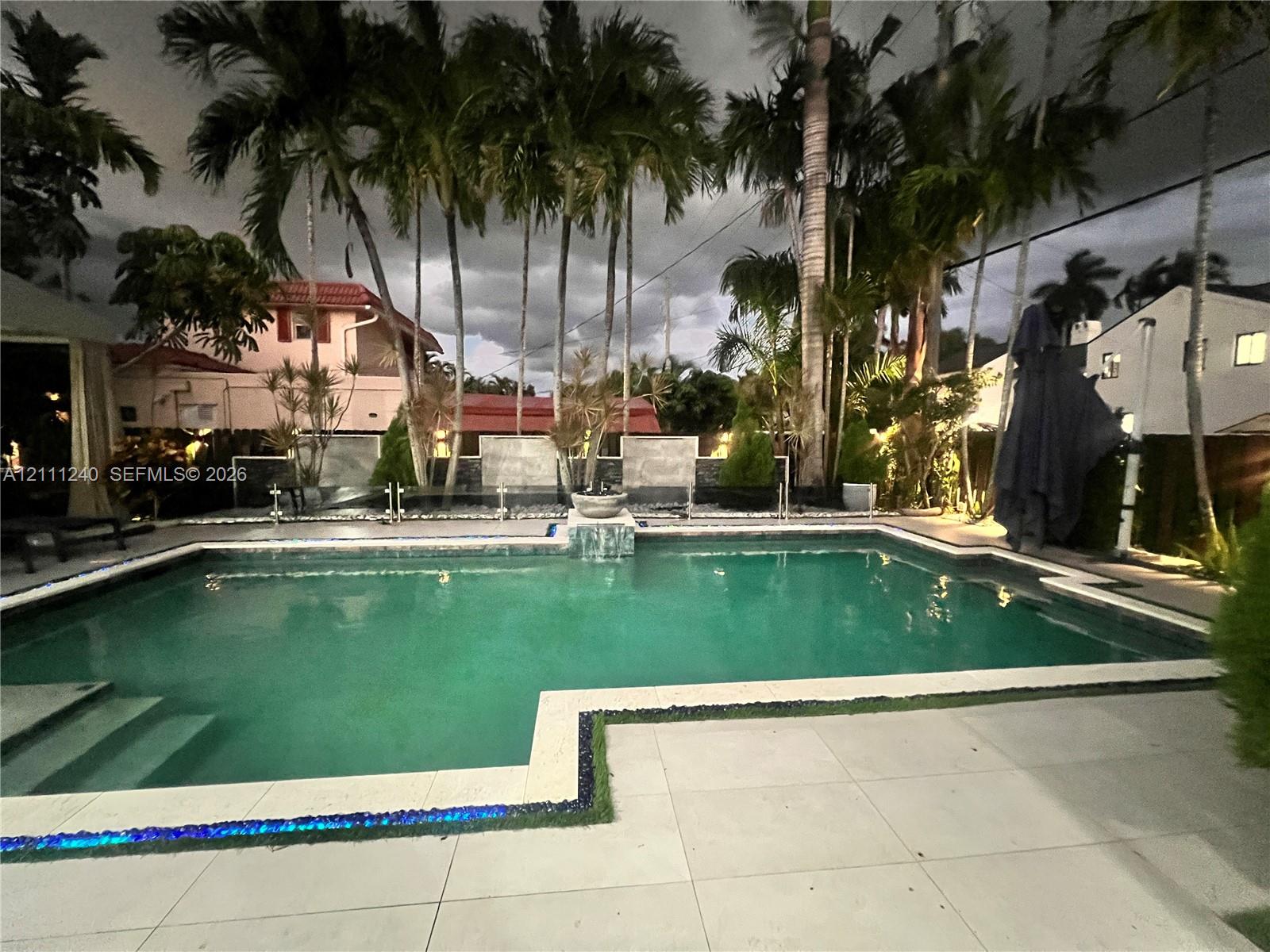 In-ground pool featuring a tiled waterline, stone-finish pool deck, glass railing, and a raised water feature