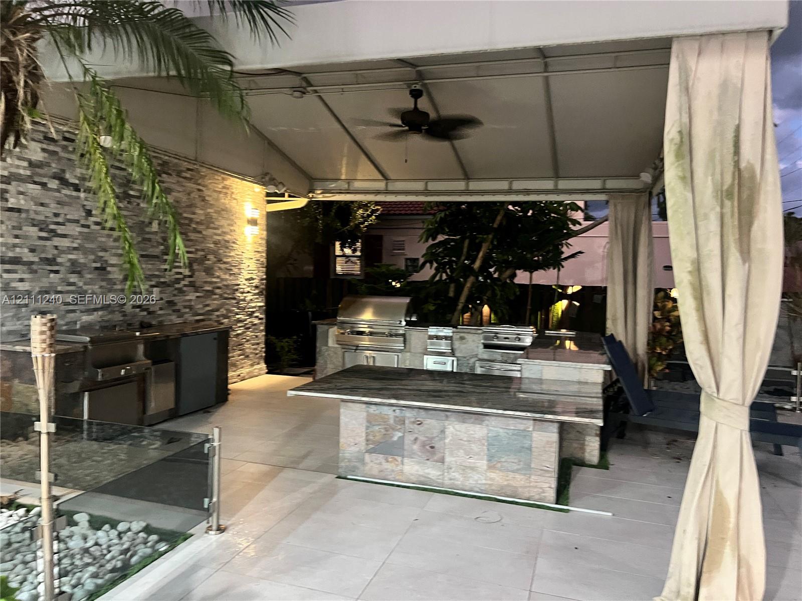 Covered outdoor kitchen featuring a stacked stone backsplash, stainless steel grill, stone-clad island with a natural stone countertop, and light-colored tile flooring