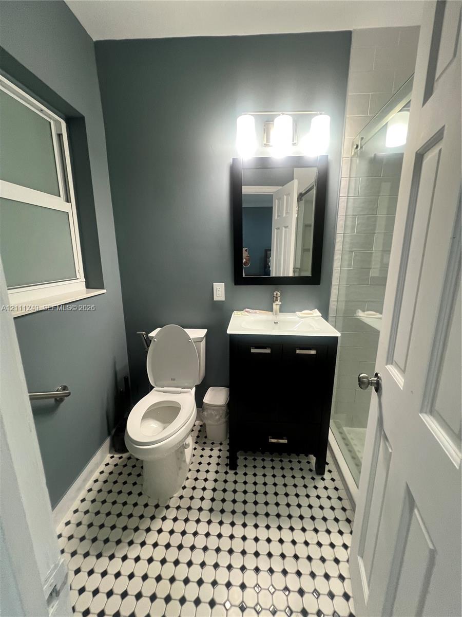 Bathroom featuring black-and-white geometric tile flooring, dark-finish vanity with a white countertop, wall-mounted mirror, and a glass-enclosed shower