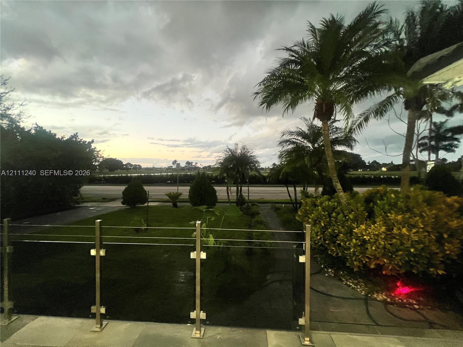 Elevated outdoor terrace featuring a stainless steel and cable railing system, stone flooring, mature palm trees, manicured landscaping, and expansive open-air horizon scenery