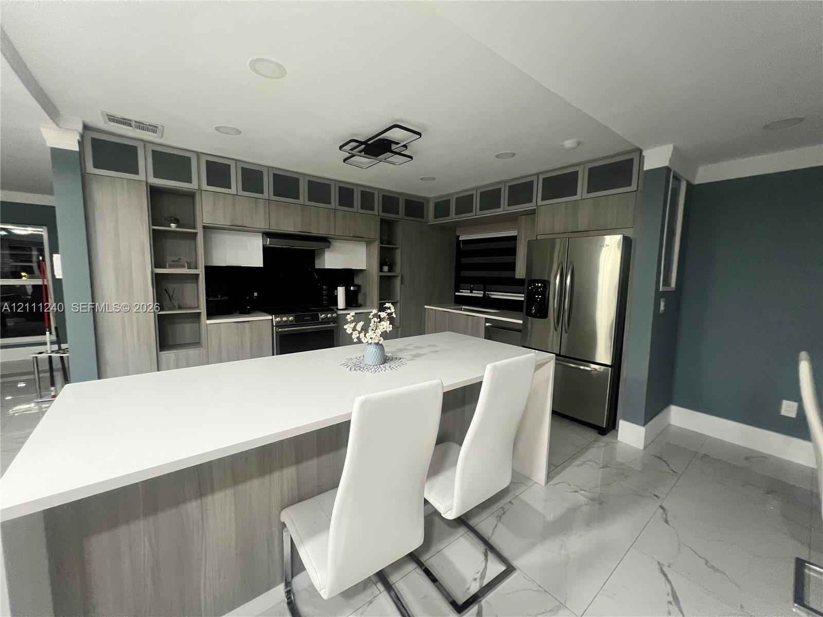 Kitchen featuring wood-finish cabinetry, floor-to-ceiling upper storage with glass-panel doors, expansive quartz-topped island, stainless steel refrigerator, and large-format porcelain tile flooring