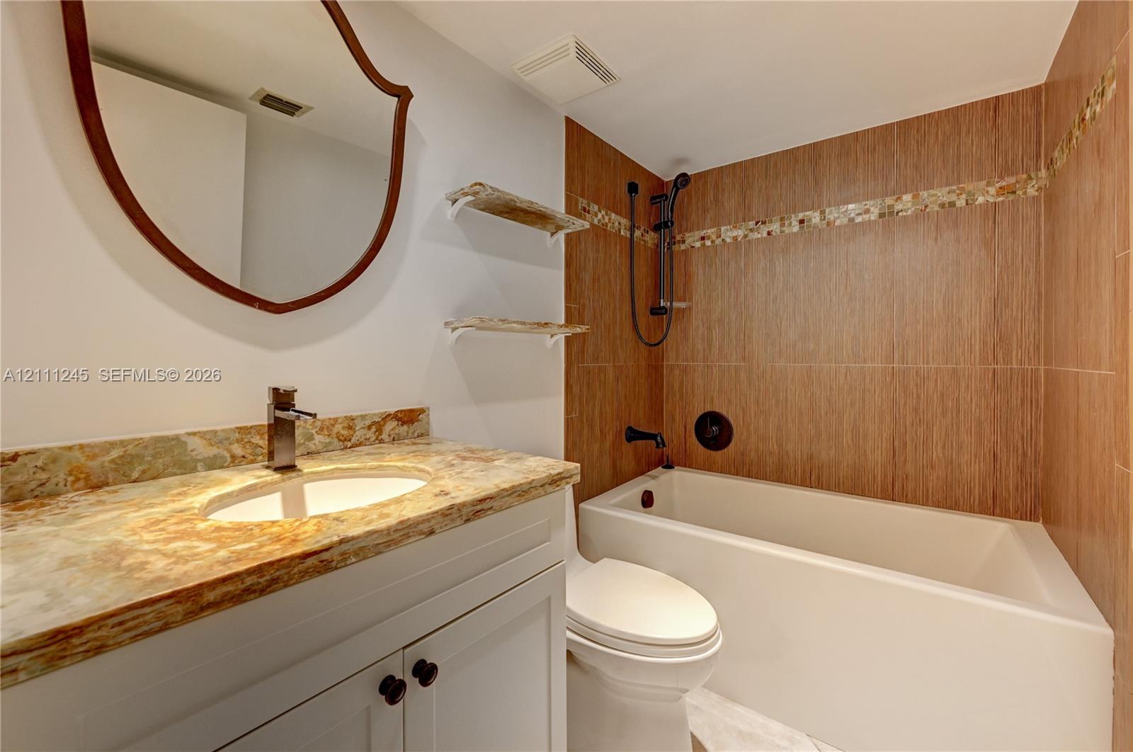 Bathroom vanity featuring a stone countertop with integrated sink, wood-finish wall tile surrounding the bathtub, matte black fixtures, and a wall-mounted mirror