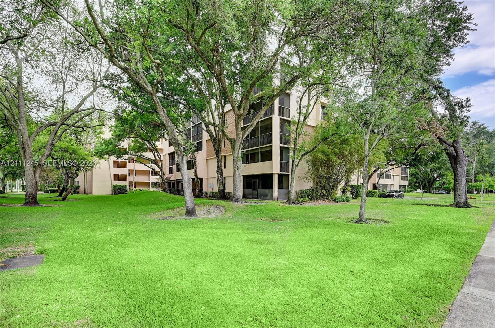Multi-story building exterior featuring screened balconies, stucco siding, and surrounding mature trees over a landscaped lawn