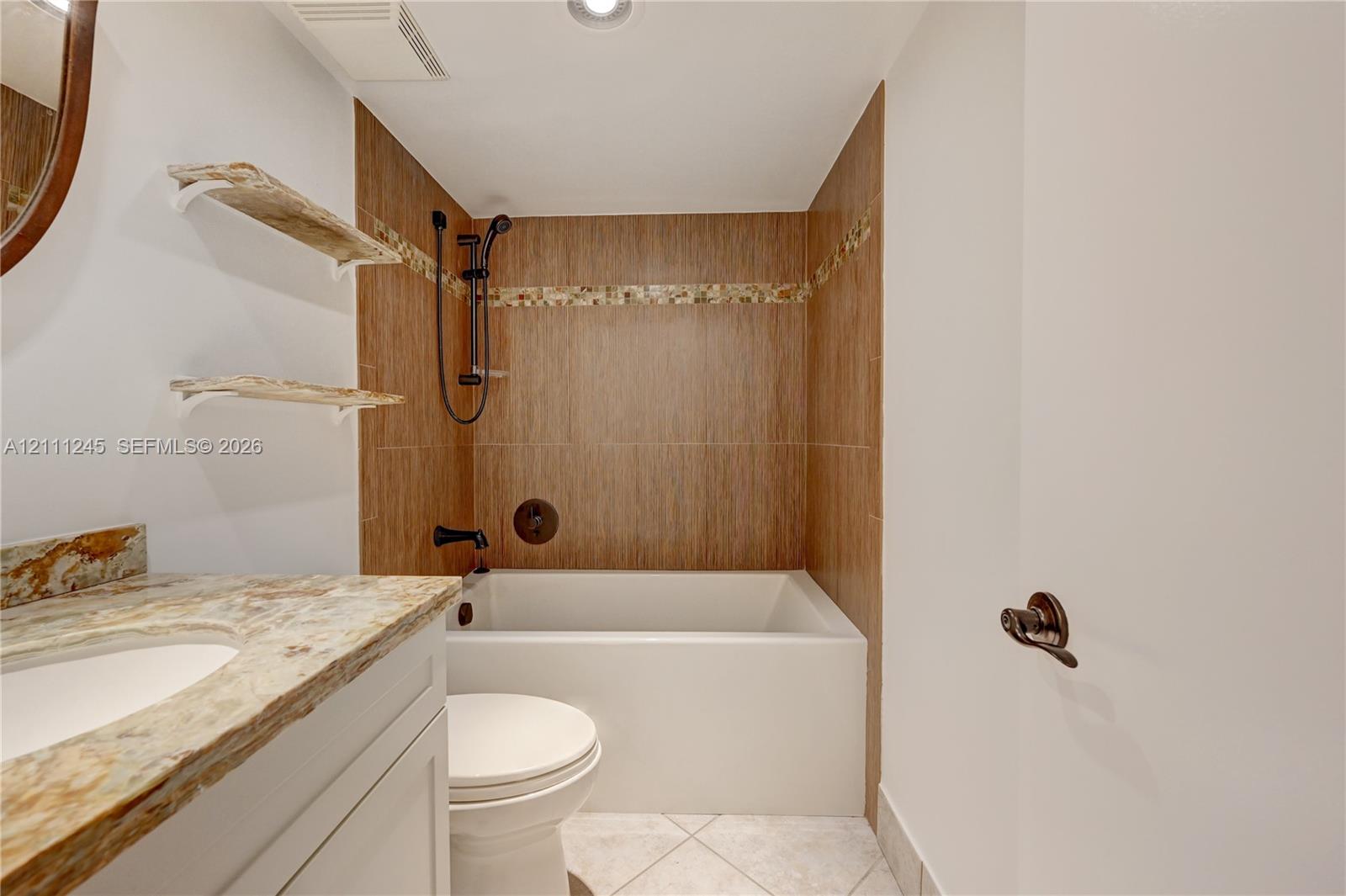 Bathroom featuring a vanity with a natural stone countertop, wall-mounted floating wood shelves, and a deep-soaking bathtub with wood-finish tile surround and matte black fixtures
