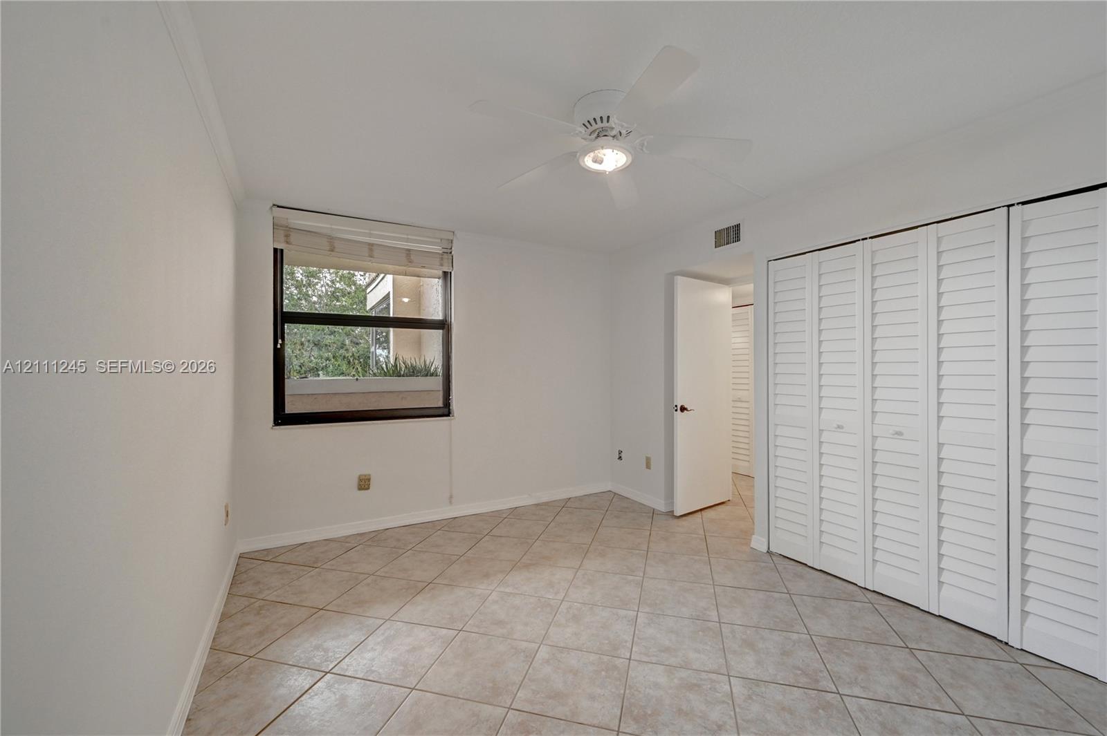 Neutral-toned tile flooring, white shutter-style closet doors, single-window illumination, and a ceiling-mounted fan with integrated lighting