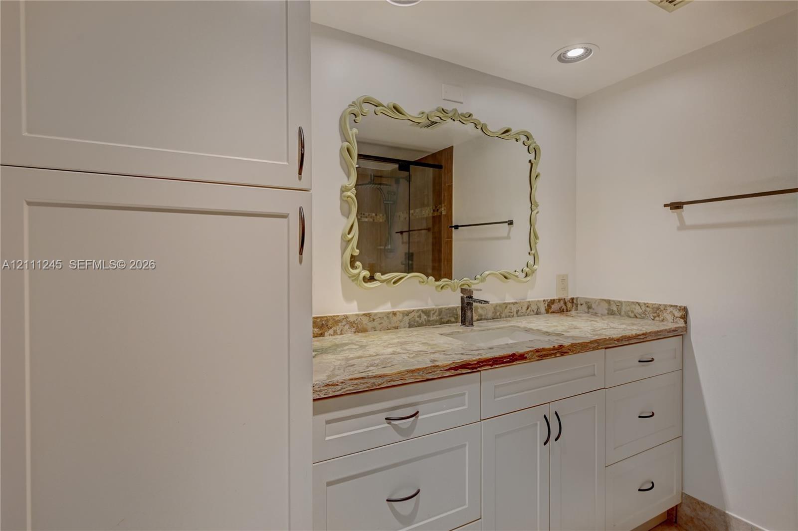 Bathroom vanity featuring white shaker-style cabinetry, multi-tonal stone countertop with an integrated sink, decorative framed wall mirror, and recessed ceiling lighting