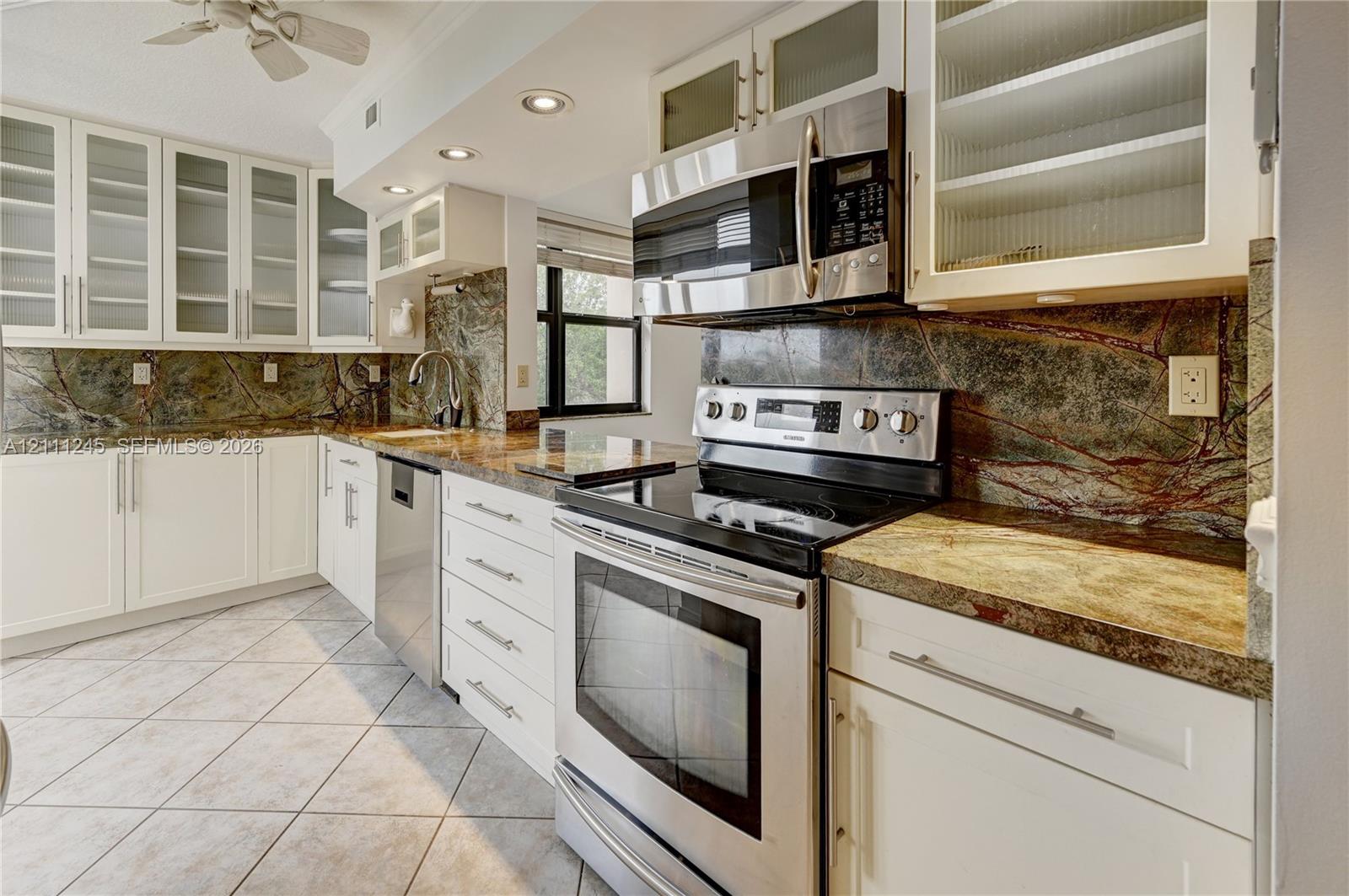 Kitchen featuring white shaker-style cabinetry with glass-front uppers, natural stone countertops, a continuous stone backsplash, stainless steel appliances, and diagonal tile flooring