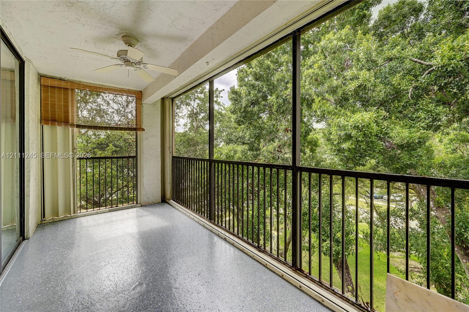 Screened balcony featuring black metal railings, textured concrete flooring, and a ceiling-mounted fan