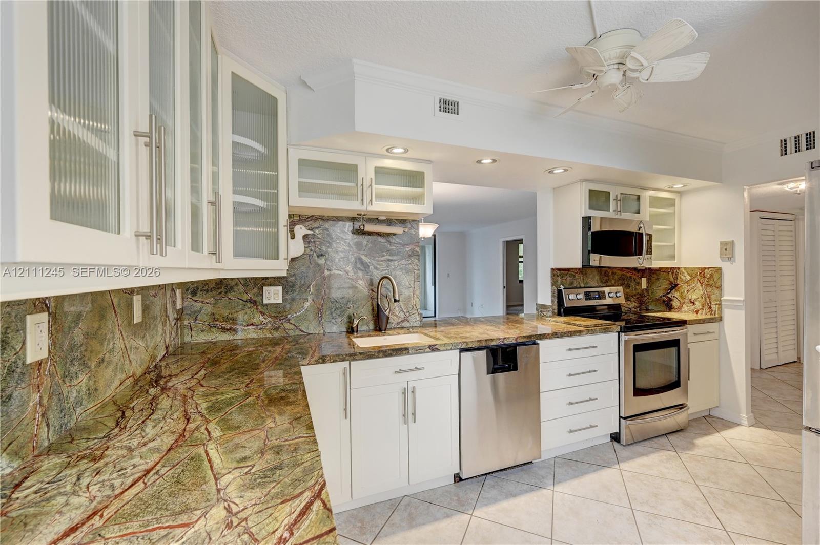 Kitchen featuring white shaker-style cabinetry with glass-front uppers, natural stone countertops, a full-height stone backsplash, stainless steel appliances, and tile flooring