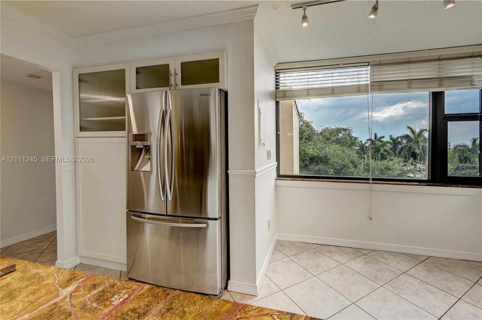 Kitchen featuring built-in cabinetry with glass-front upper panels, stainless steel French-door refrigerator, large-format tile flooring, multi-directional track lighting, and a window with exterior greenery