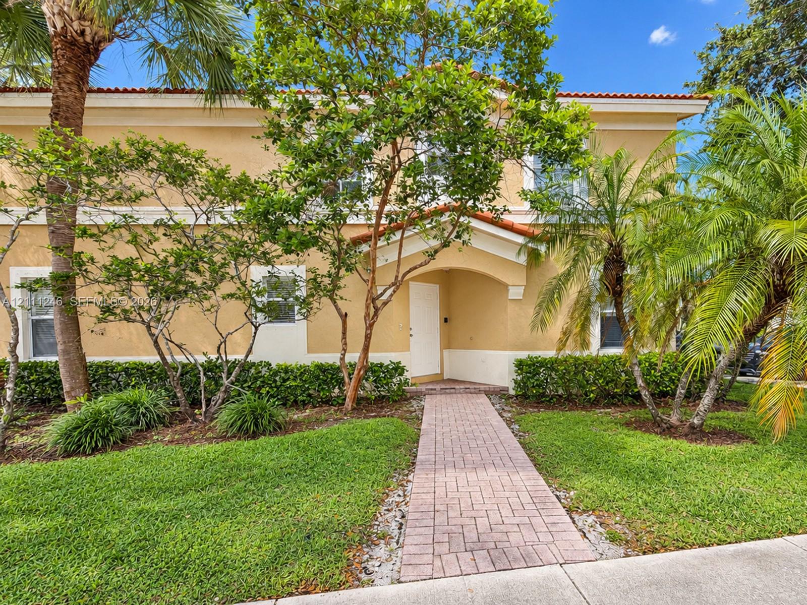 Two-story stucco exterior with a tiled roof, covered entryway, and stone paver walkway surrounded by mature landscaping