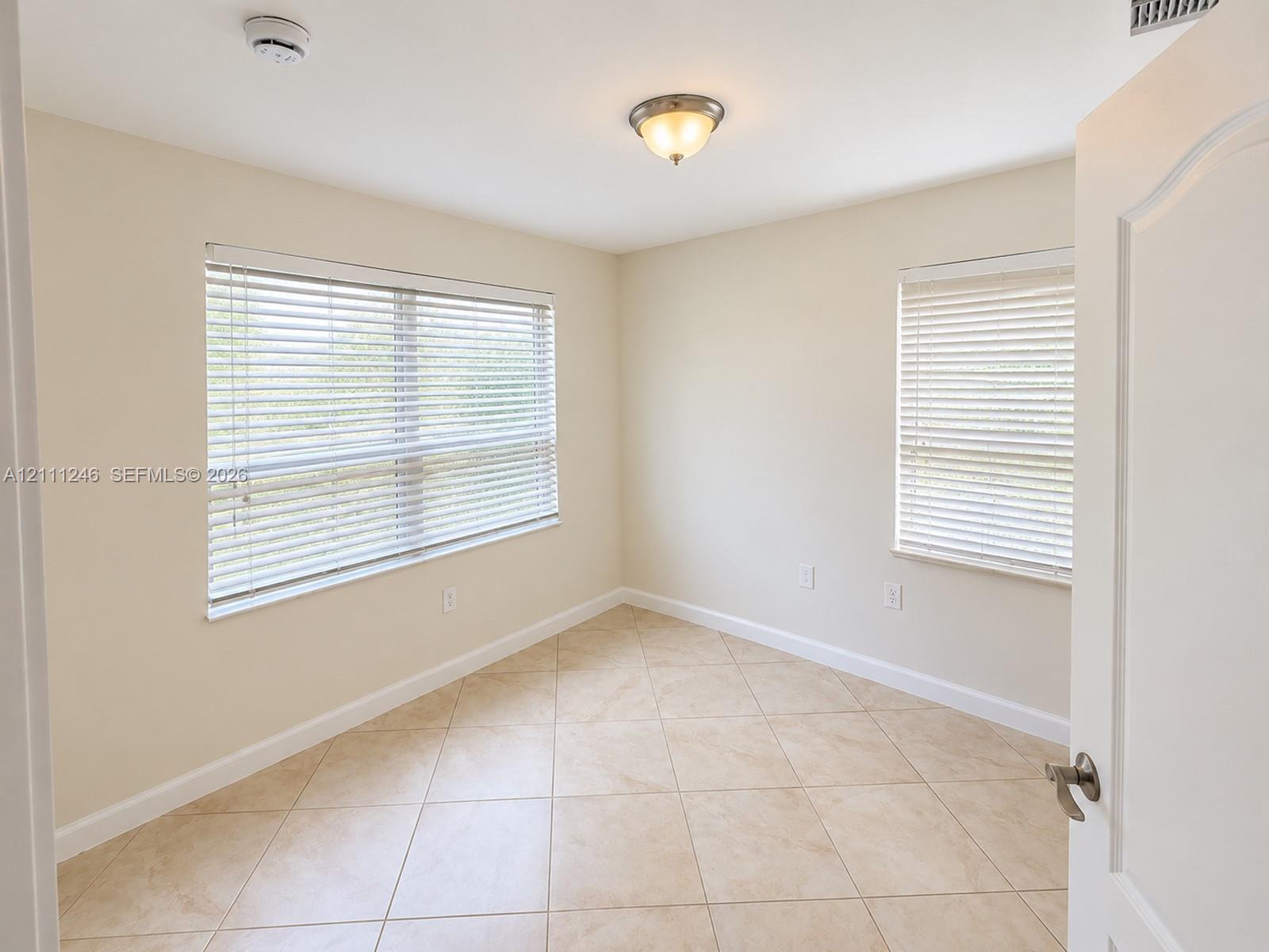 Neutral-toned room featuring diagonal tile flooring, dual windows with horizontal blinds, and a brushed nickel flush-mount ceiling light fixture