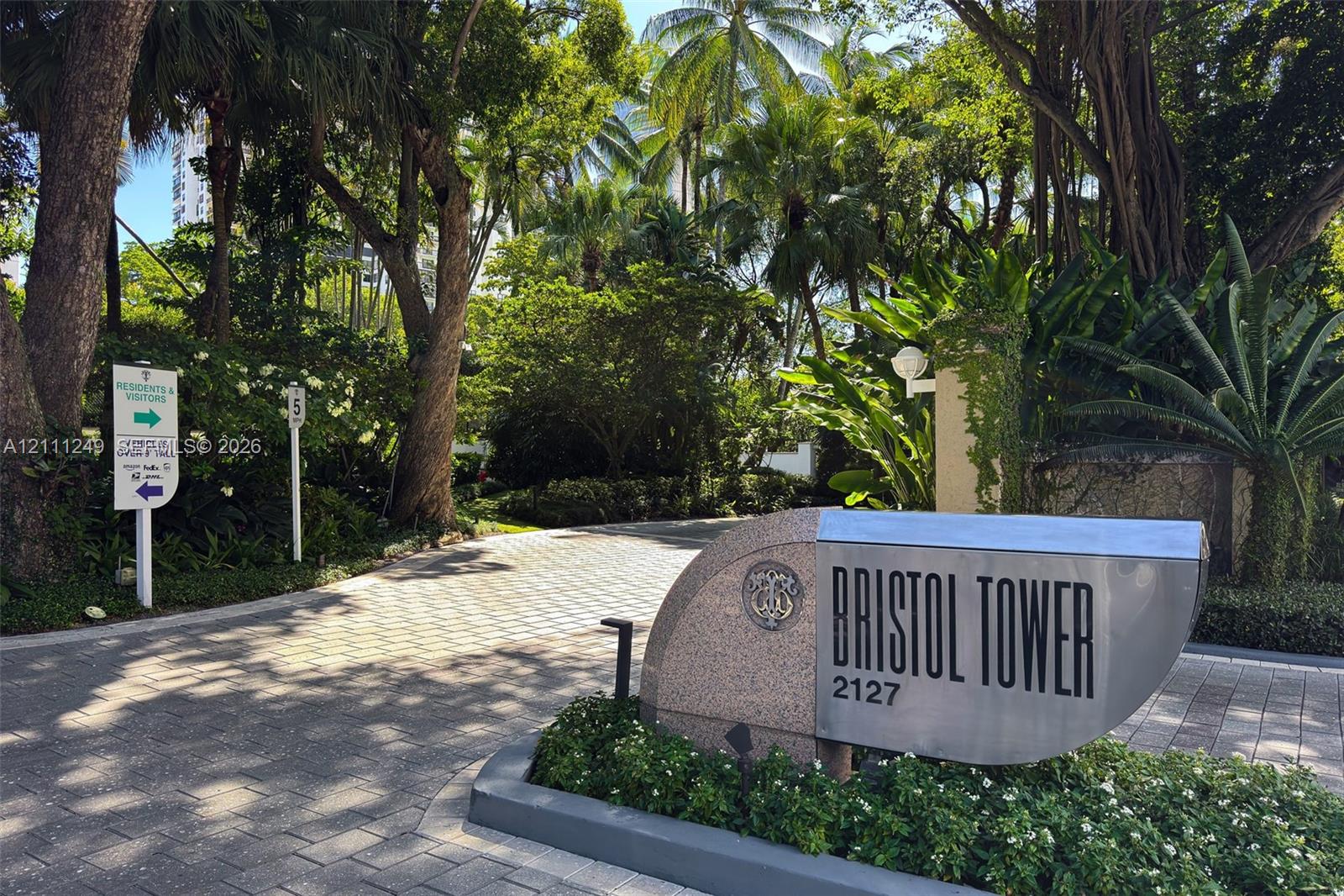 Gated property entrance featuring a masonry monument sign, paver driveway, and mature tropical landscaping