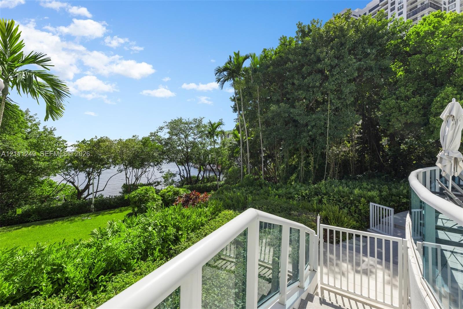 Elevated terrace featuring glass railings, a white metal gate, concrete surfacing, and mature landscaping overlooking the water