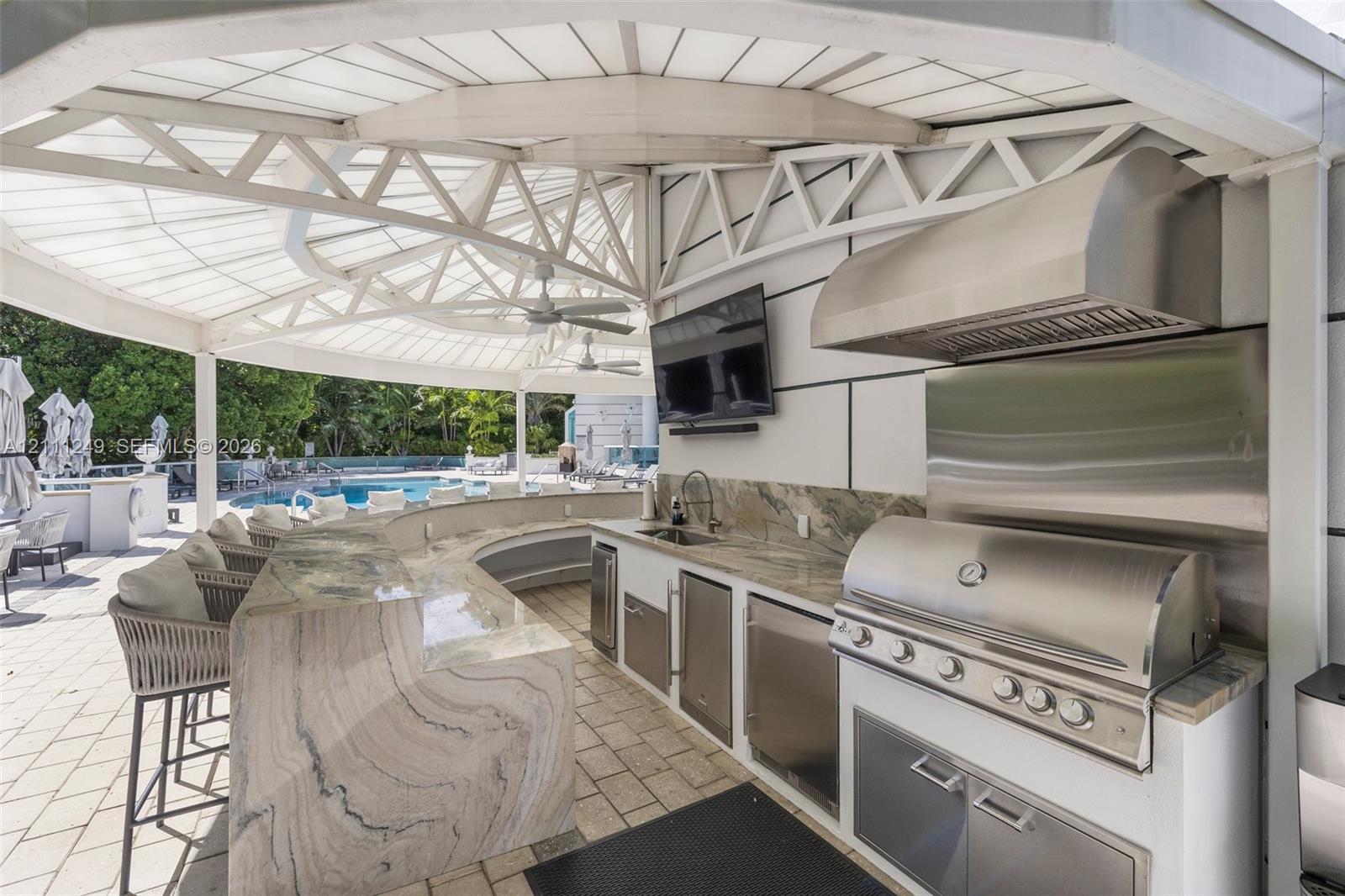 Covered outdoor kitchen featuring a stone-topped bar, stainless steel grill, professional-grade range hood, built-in sink, and custom cabinetry under a high-arched structural canopy