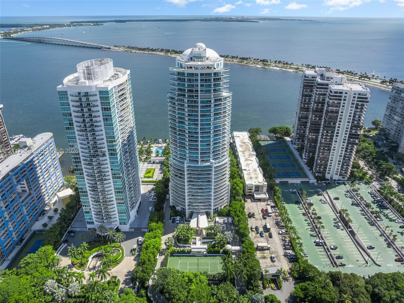 High-rise residential tower featuring glass-paneled balconies, direct coastal proximity, paved tennis courts, and landscaped grounds