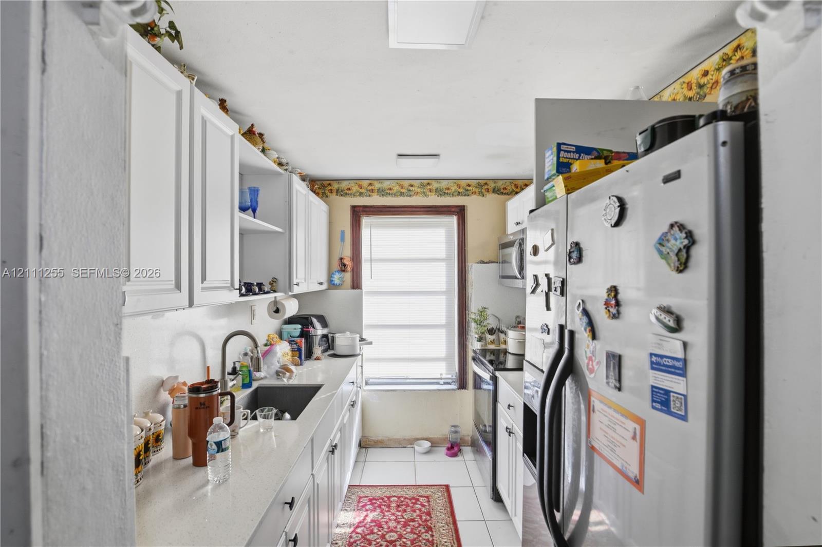 Galley kitchen featuring white cabinetry, solid-surface countertops, an undermount sink with a gooseneck faucet, tile flooring, and a window with wood trim