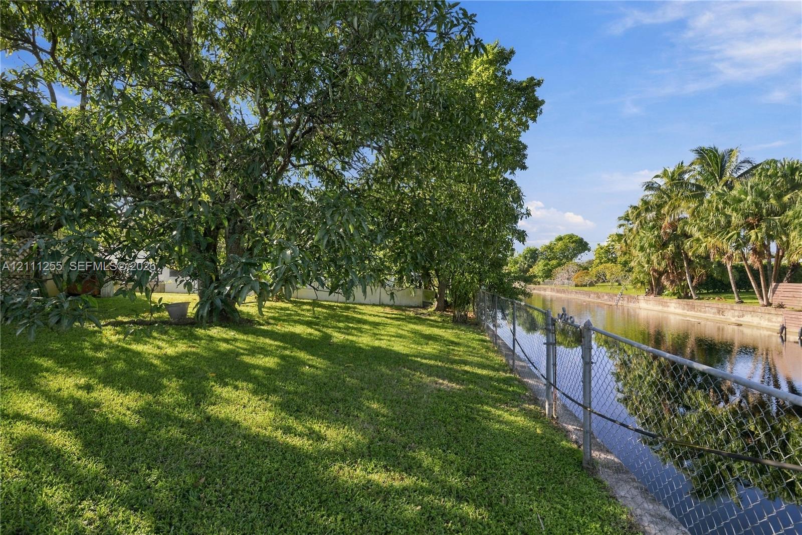 Waterfront backyard featuring a manicured lawn, mature trees, and chain-link perimeter fencing
