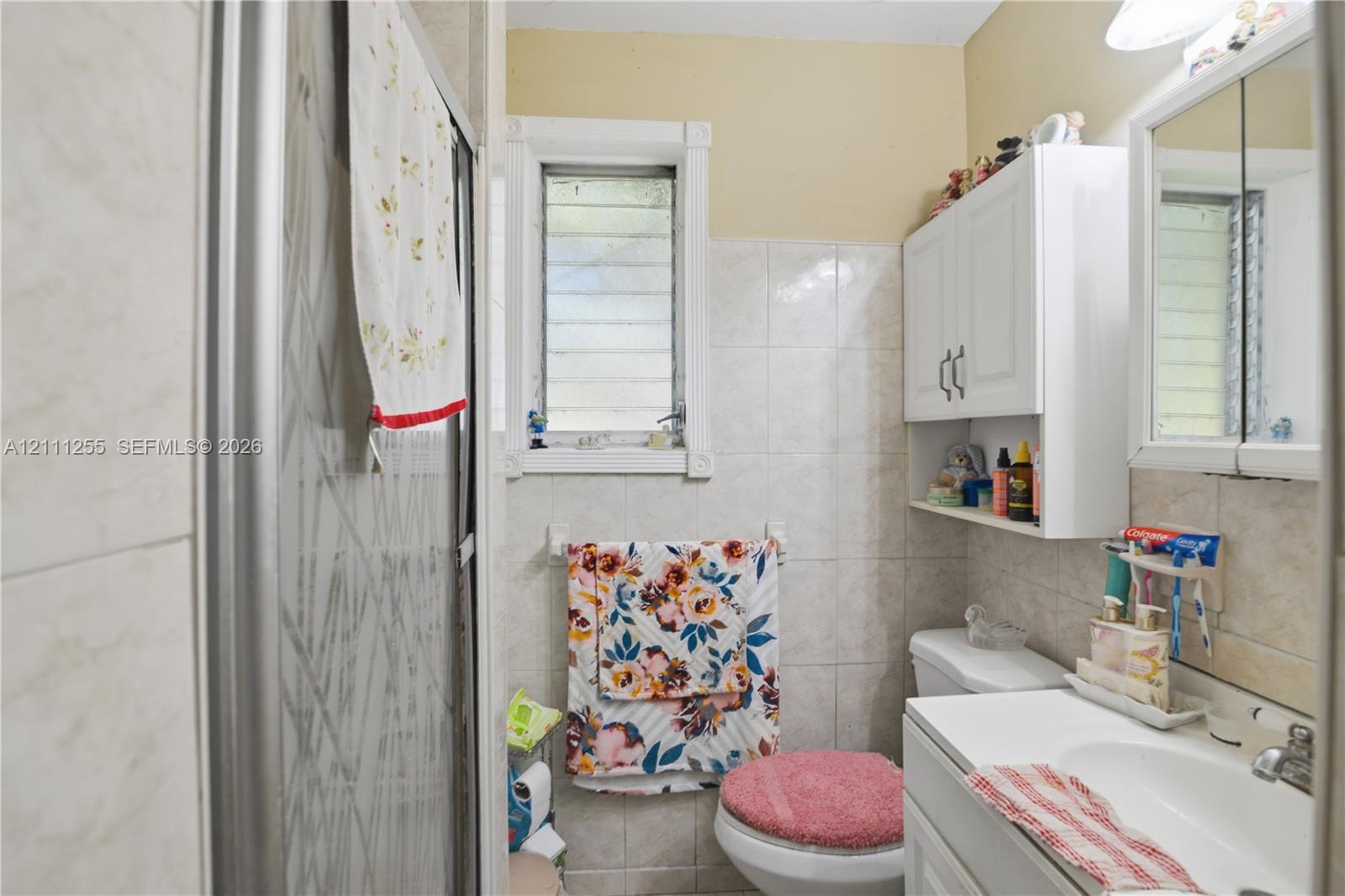 Full-height wall tiling, glass-enclosed shower stall, white vanity cabinet with integrated sink, and upper wall-mounted cabinetry