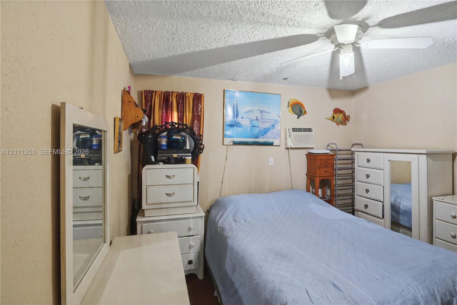 Room featuring a textured ceiling, a central white ceiling fan with integrated lighting, and a wall-mounted air conditioning unit
