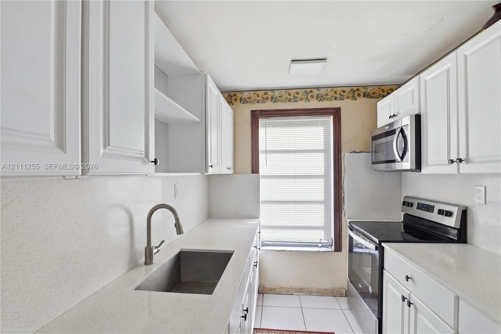 White cabinetry, solid surface countertops, stainless steel undermount sink with a gooseneck faucet, stainless steel microwave, and white tile flooring