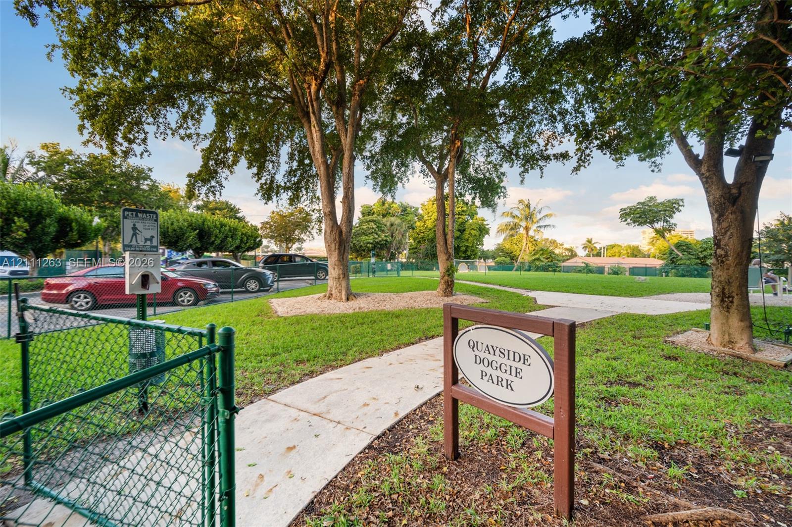 Enclosed recreation area featuring green space, concrete pathways, mature trees, and chain-link fencing
